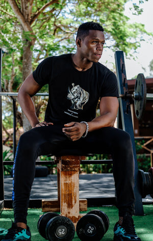 men's comfortable gym top, sustainable gift, Sitting on gym bench weights below wearing black rhino tee and black sweatpants. Top has print of black rhino designed by tanzanian artist.